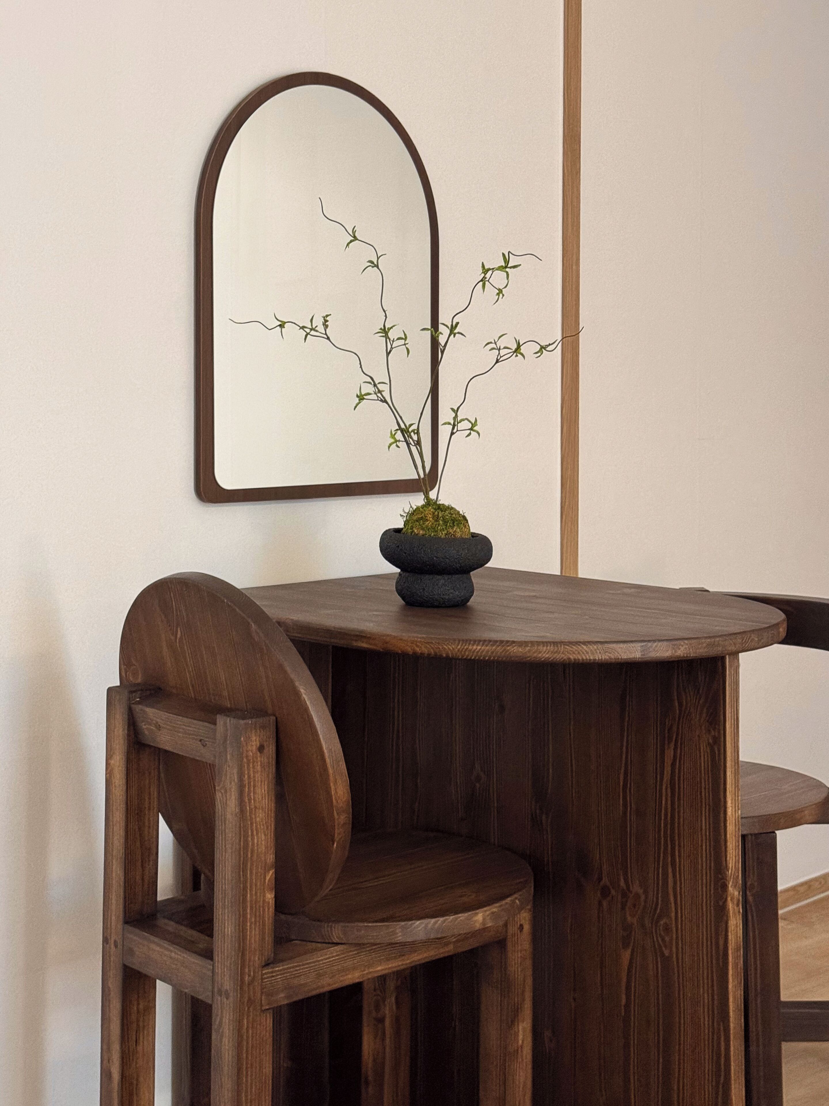 Una mesa redonda de madera con una planta en maceta, una silla de madera y un espejo fijado a la pared.