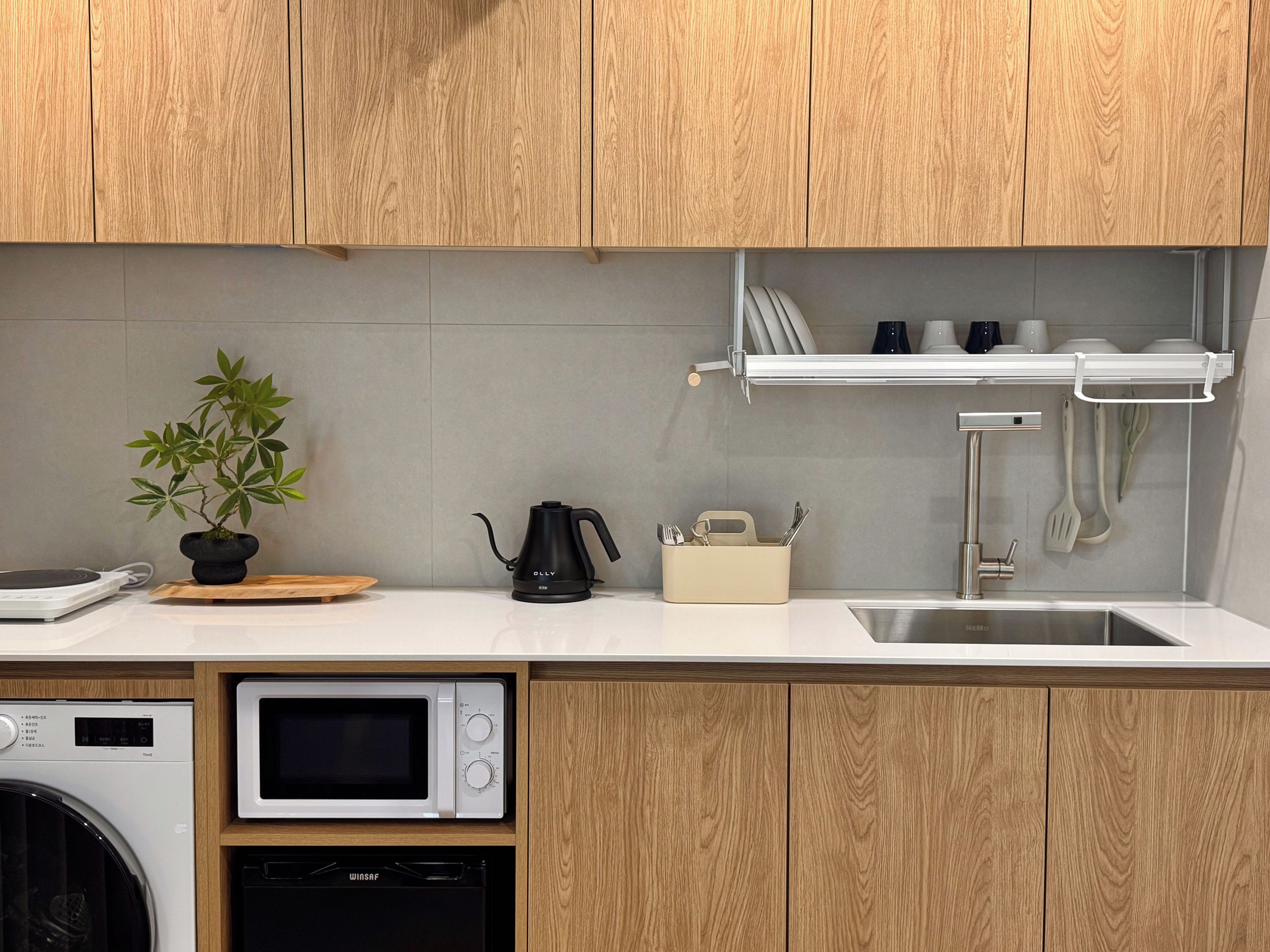 A kitchen with wooden cabinets, a microwave, a dishwasher, a kettle, and a shelf with dishes and utensils.