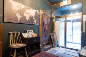 Um quarto com um mapa-múndi na parede, uma cadeira de madeira, uma mesinha com uma placa e uma porta que dá para o exterior.