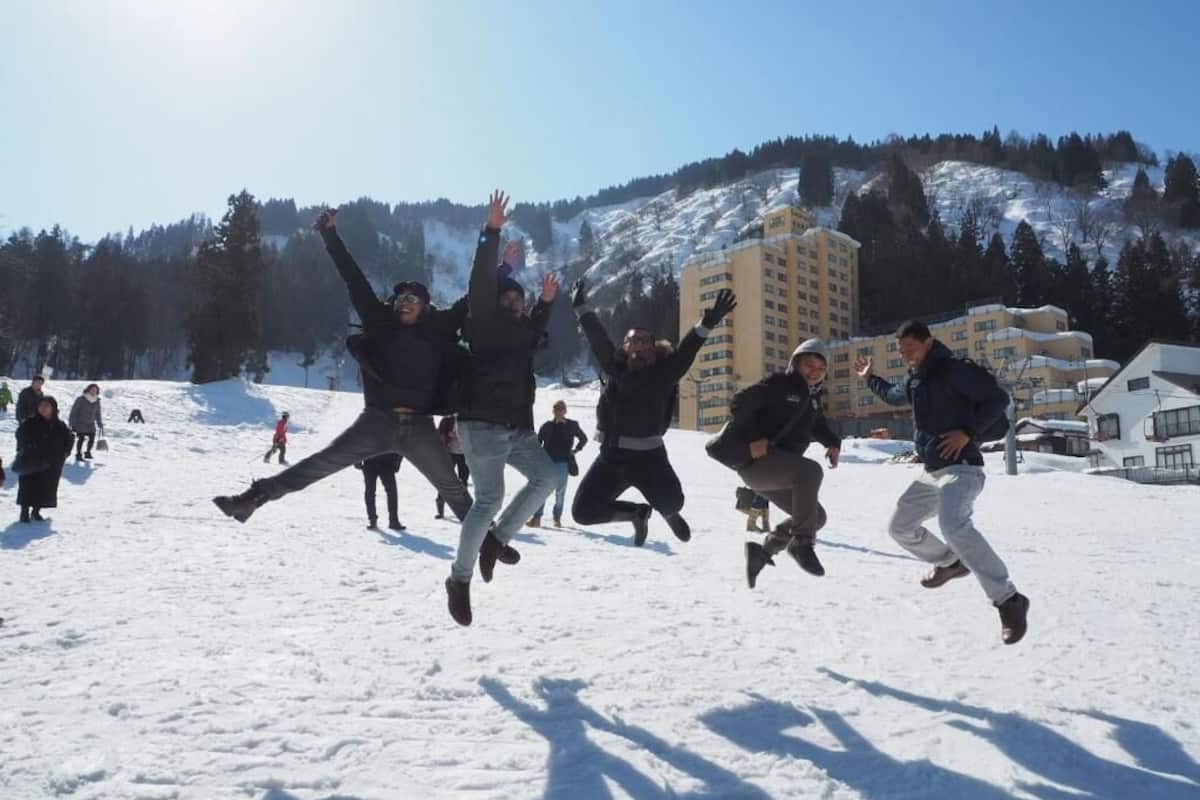 A group of people jumping in a snowy outdoor area with buildings in the background.