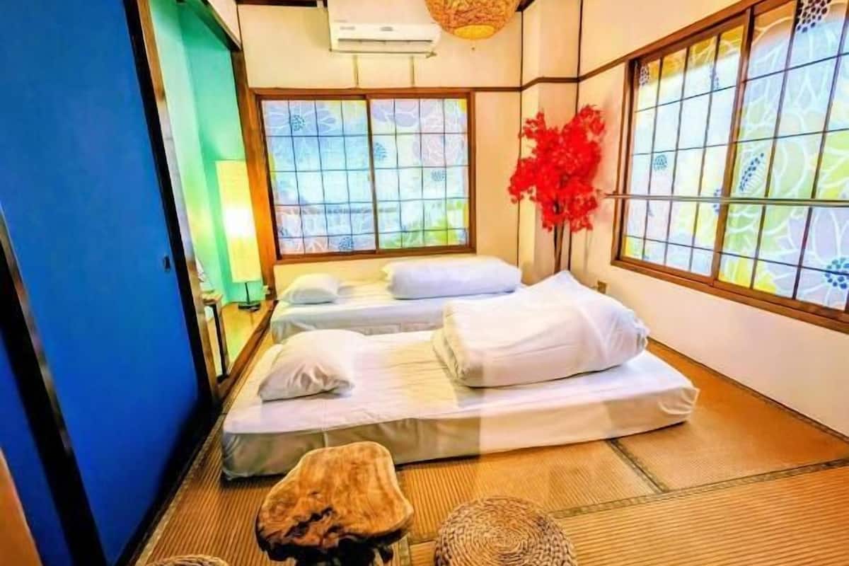 A traditional Japanese-style room with tatami flooring, a bed with white bedding, a hanging light fixture, and a window with a view.