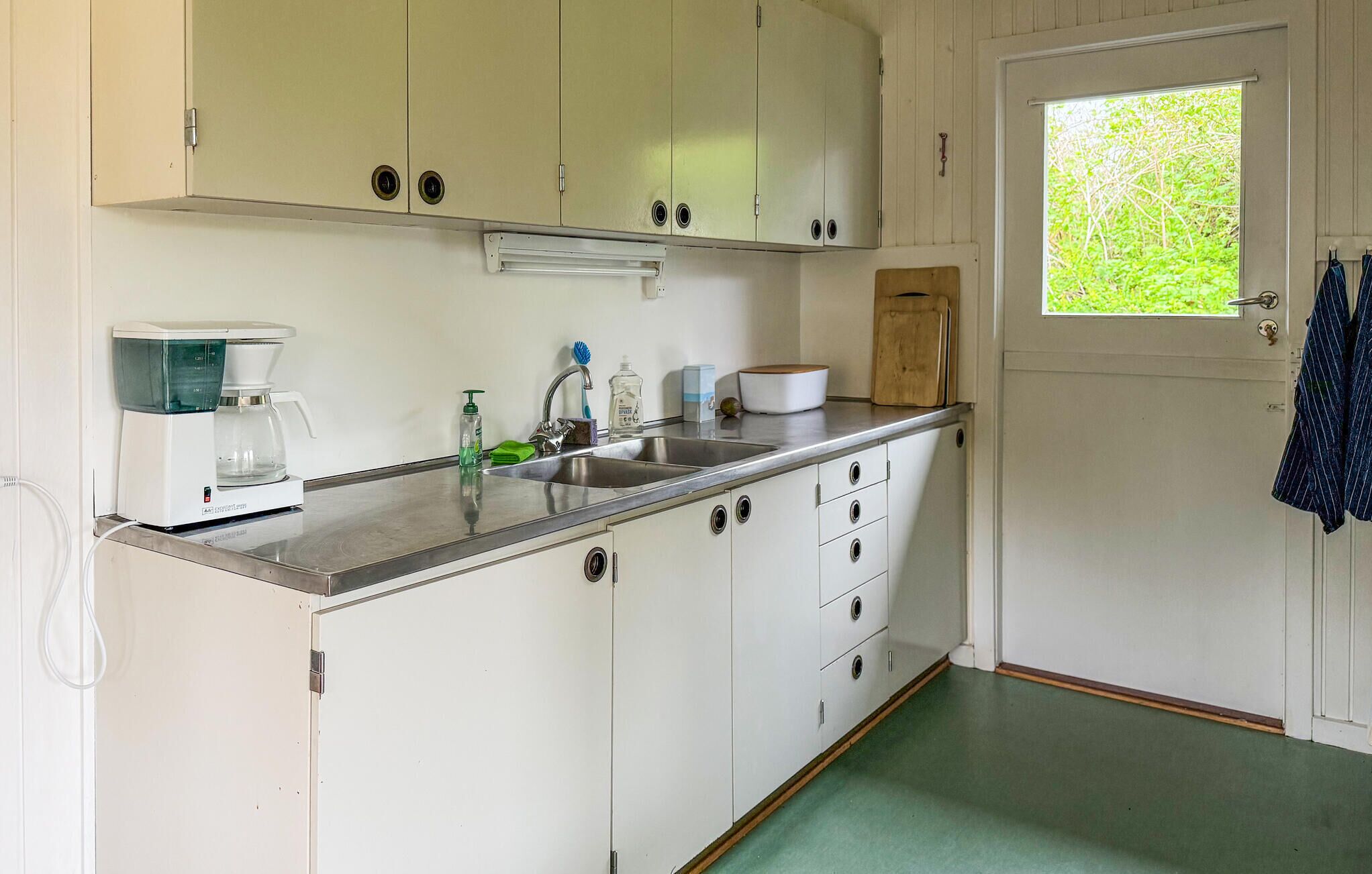A kitchen with white cabinets, a coffee maker, a sink, and a window with a view of greenery.