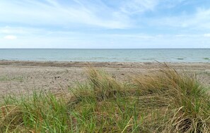 Een strand met zand en weinig gras op de voorgrond, dat uitloopt in een kalme zee onder een lichtbewolkte lucht.