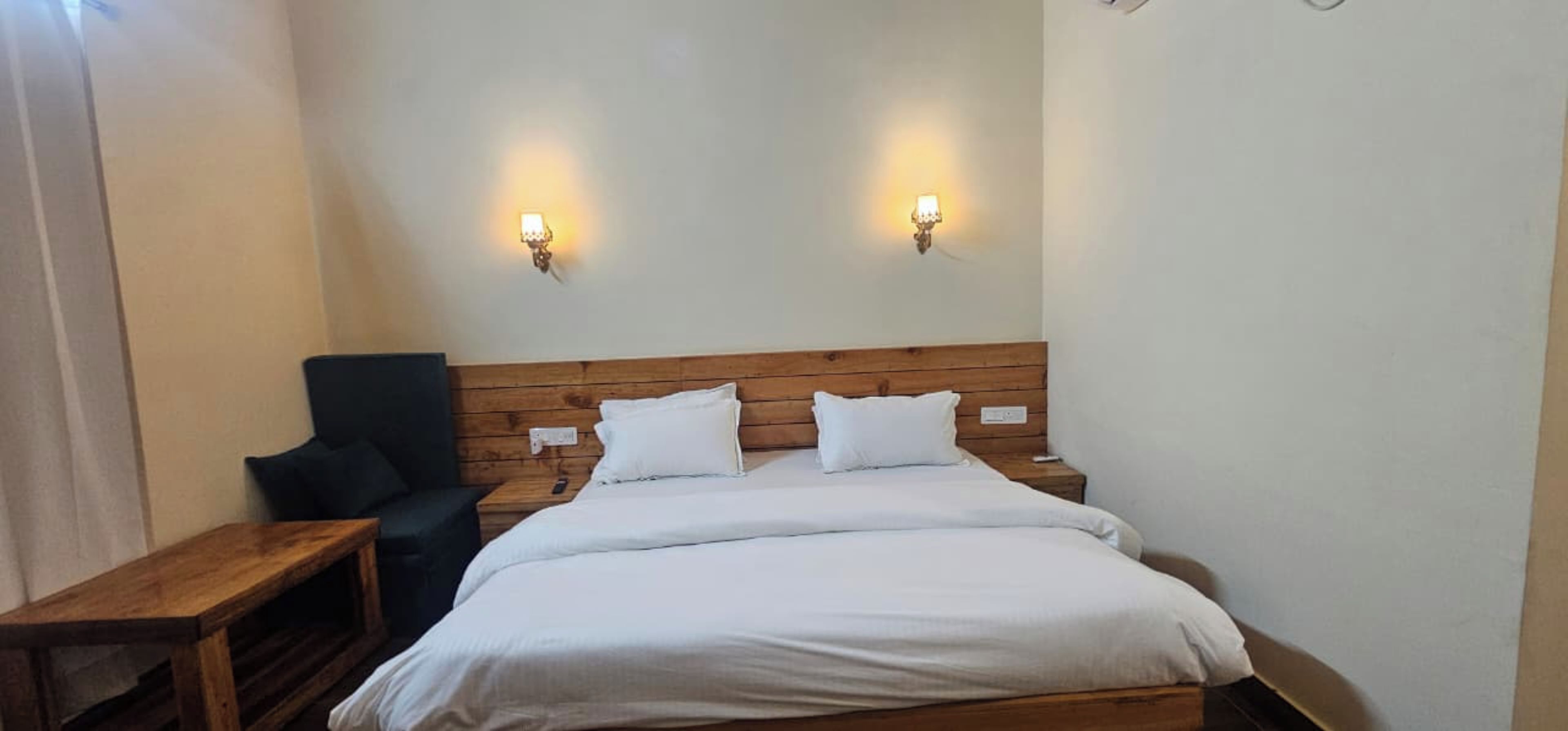 Habitación de hotel con cabecera de madera, cama individual con ropa de cama blanca, mesita de noche de madera, sillón negro y dos lámparas de pared.