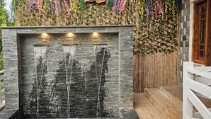 Um elemento decorativo de pedra com três bicas, uma cerca de madeira e um muro com plantas pendentes.