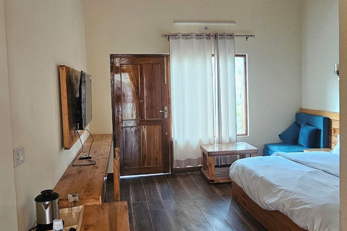 Habitació amb sostre de fusta, un llit, escriptori amb tetera, televisió i finestra amb cortines.