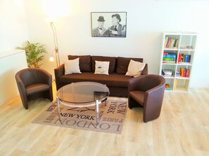 Ein Wohnzimmer mit braunem Sofa, zwei Sesseln, einem Glastisch, einem Bücherregal und einem gerahmten Bild an der Wand.