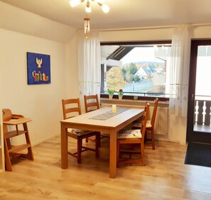 A dining area with a wooden table and chairs, a ceiling light fixture, a window with curtains, and a painting on the wall. - Vacation apartment Anna (Winterberg)