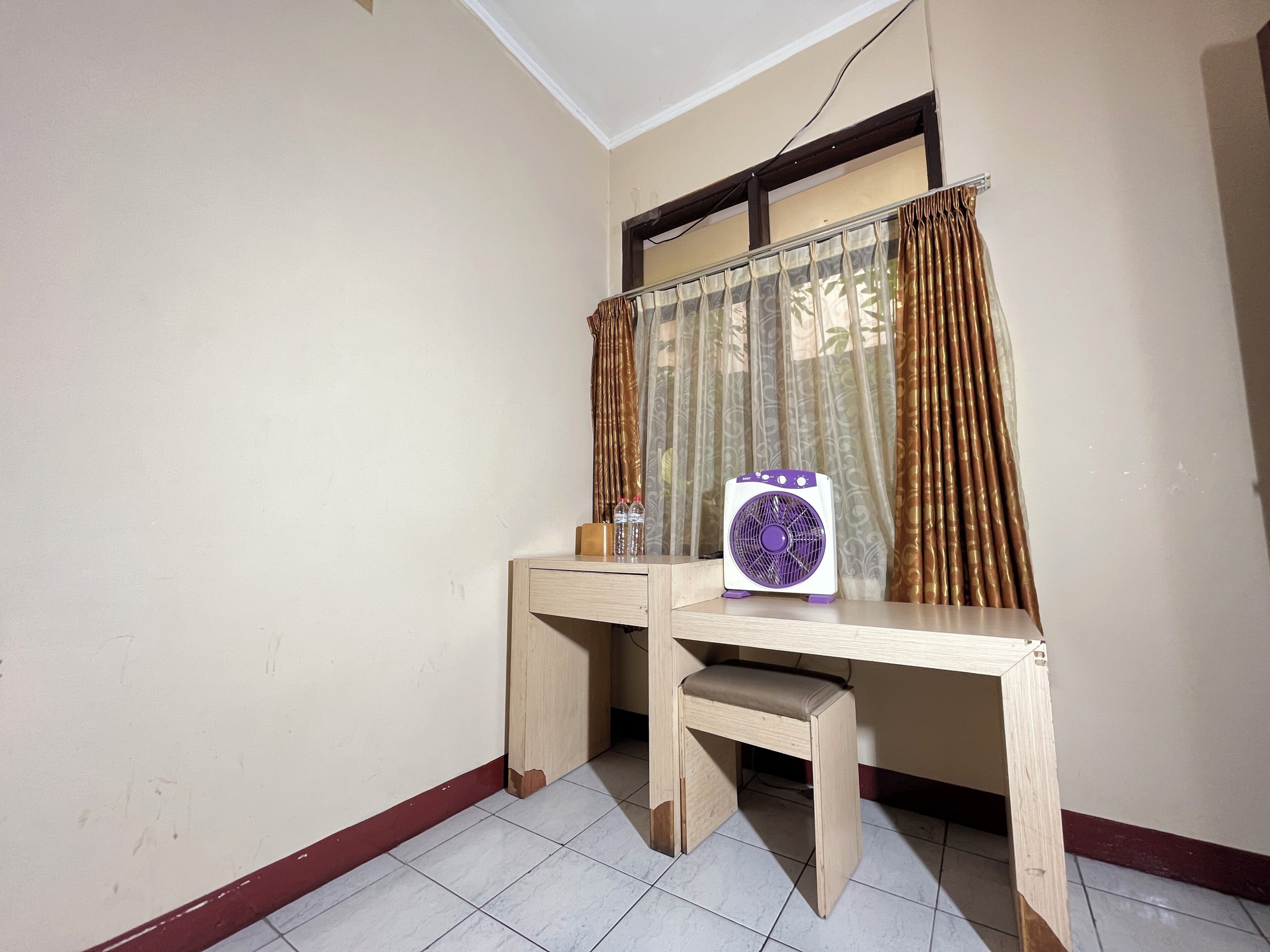 A corner room with a desk, a chair, a fan, and a window with curtains.