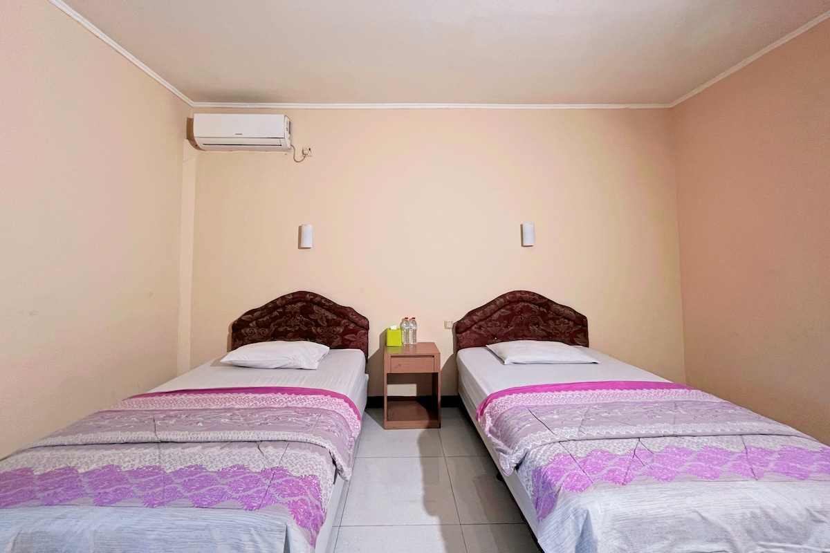 Una habitación pequeña y limpia con dos camas individuales, una mesita de noche, un aire acondicionado de pared y una pequeña mesilla con una lámpara.