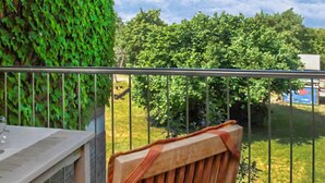 Ein Balkon mit Metallgeländer, einem Holzstuhl und Blick auf grüne Bäume sowie ein Gebäude im Hintergrund.