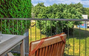 Ein Balkon mit Metallgeländer, einem Holzstuhl und Blick auf grüne Bäume sowie ein Gebäude im Hintergrund.