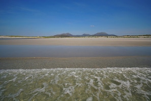 A beach with calm waters, distant mountains, and a clear sky.