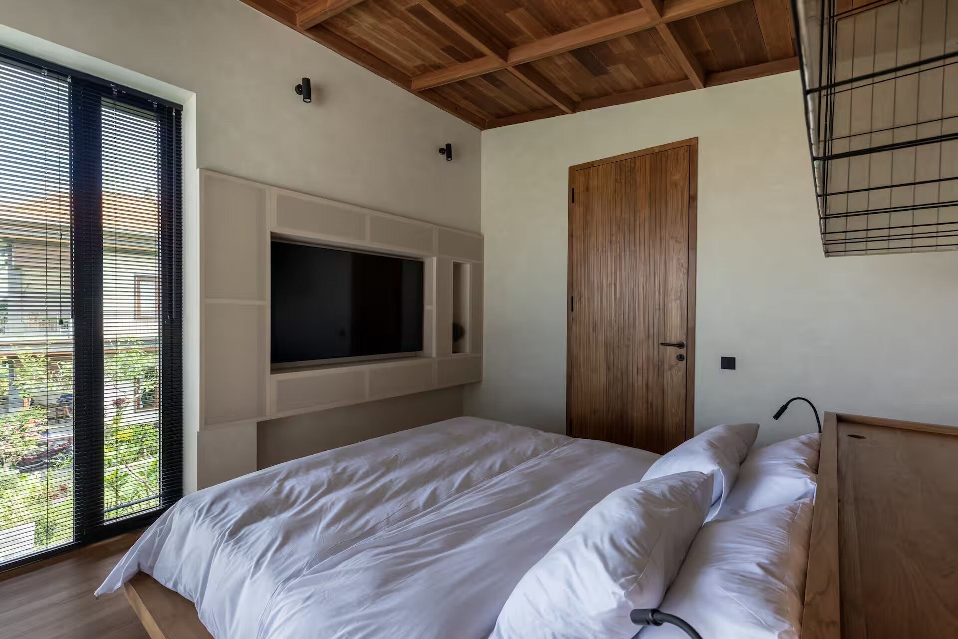 A bedroom with a bed, a TV mounted on the wall, a wooden door, and a window with blinds.