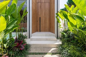A modern entrance with a wooden door, concrete steps, and lush tropical plants. - Art Villas (Badung)