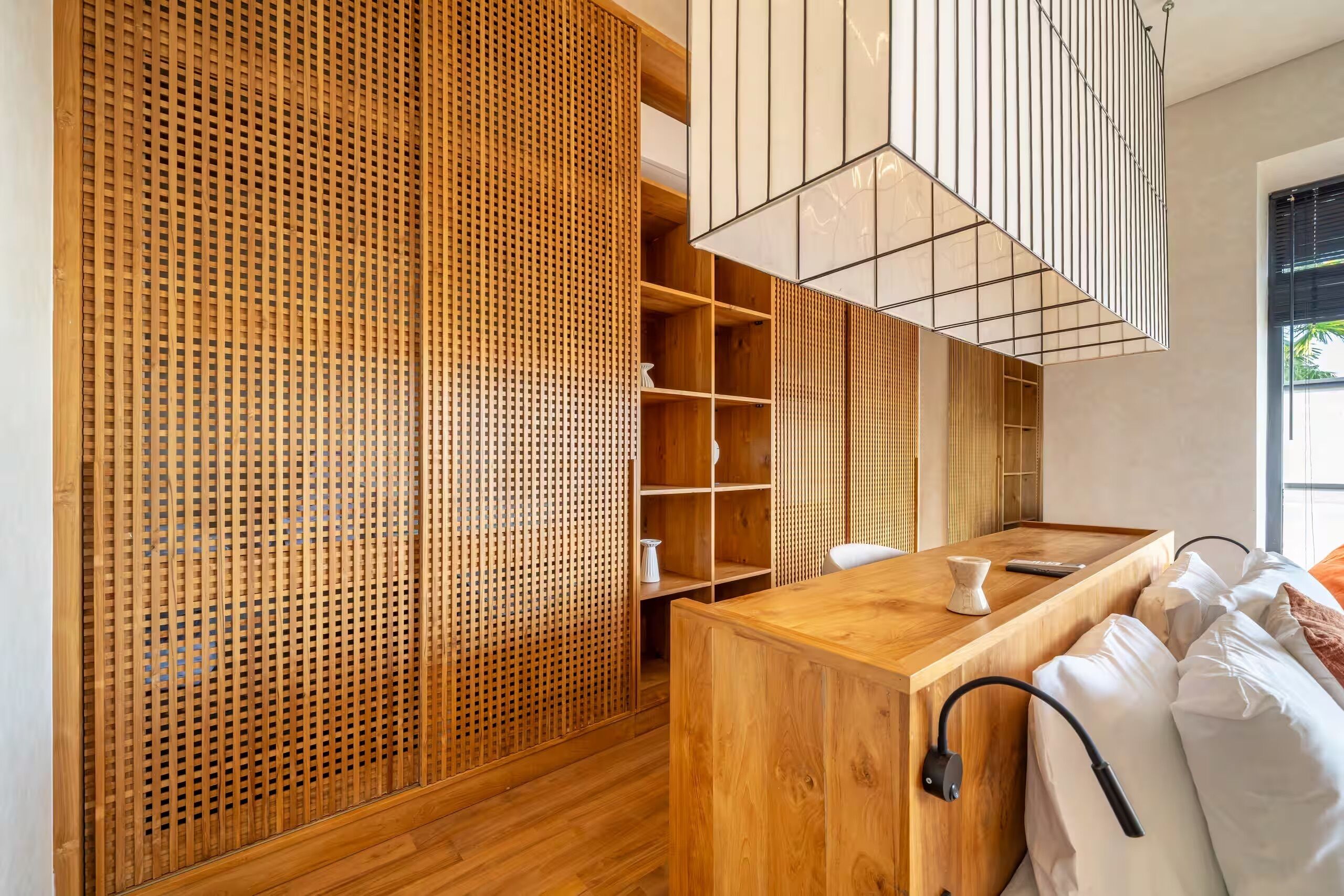 A modern hotel room with a wooden floor, a wooden desk, a bed with white bedding, a wooden partition with a lattice design, and a hanging light fixture.
