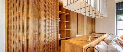 A modern hotel room with a wooden floor, a wooden desk, a bed with white bedding, a wooden partition with a lattice design, and a hanging light fixture.
