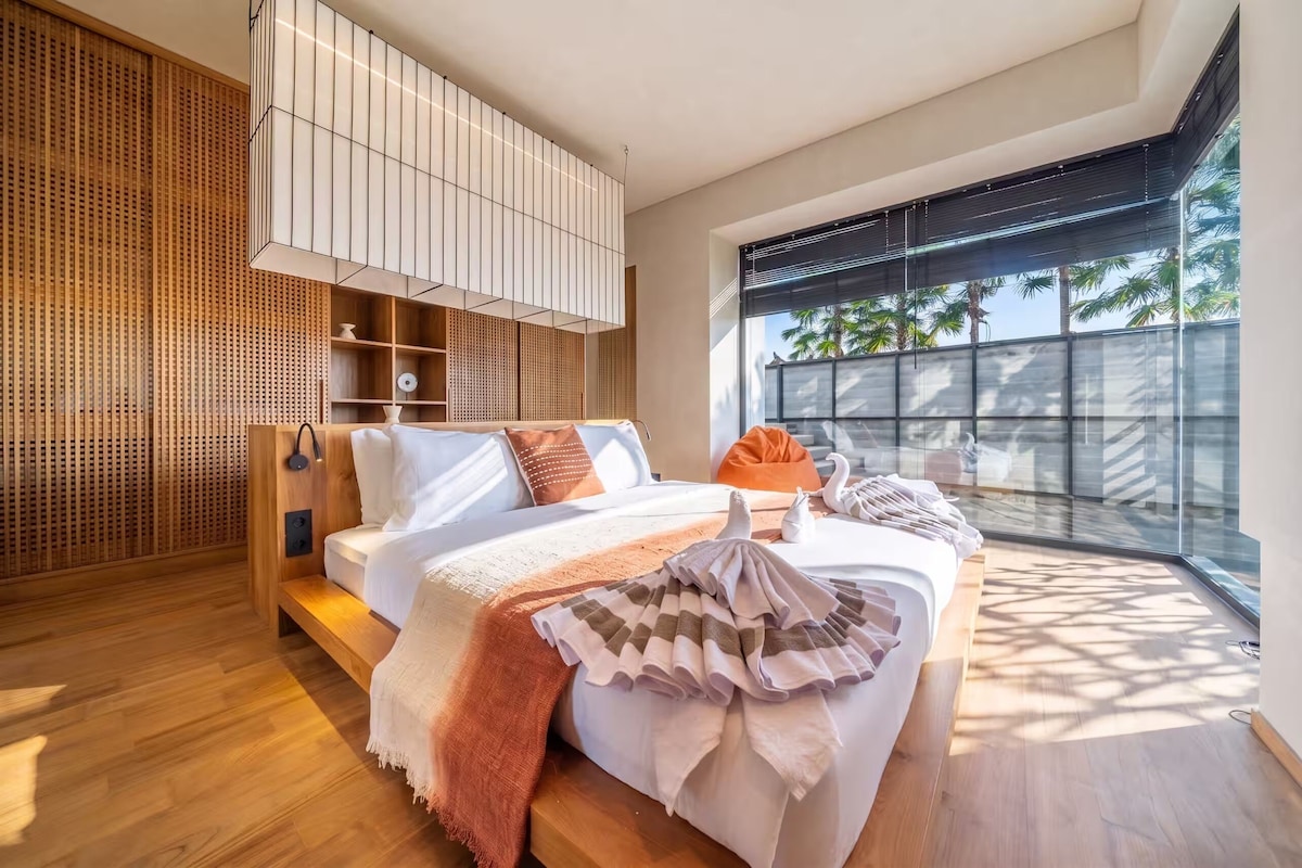 A modern bedroom with a bed, wooden floor, and a large window with a view of palm trees.