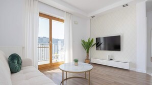 Ein modernes Wohnzimmer mit einer Couch, einem runden Couchtisch, einem Blumentopf, einem flachbildfernseher an der Wand und einer Glasschiebetür, die auf einen Balkon führt.