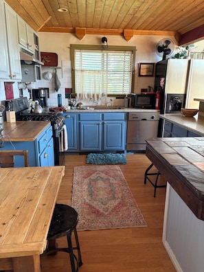 A kitchen with blue cabinets, a wooden table, a rug, and a fan.