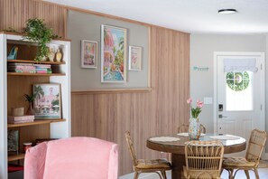Un comedor con una mesa redonda, sillas de mimbre, una estantería con libros y objetos decorativos, y obras de arte colgadas en la pared.