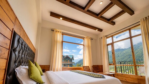 Un dormitorio con cabecera de madera, una cama grande con ropa blanca, dos ventanas con cortinas y vistas a un paisaje montañoso.