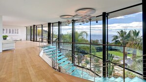 Un intérieur moderne avec un escalier en verre menant à un balcon offrant une vue sur la mer et les palmiers.