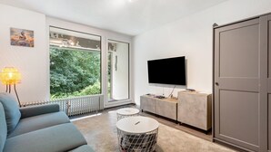 Ein modernes Wohnzimmer mit einer Couch, zwei runden Tischen, einem Fernseher und einem großen Fenster mit Blick auf Grünflächen.
