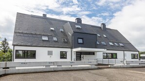 Ein modernes, zweistöckiges Gebäude mit geneigtem Dach, zahlreichen Fenstern und einer gepflasterten Einfahrt.