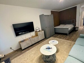 Un saló modern amb televisió de pantalla plana, armari gris, llit i dues taules rodones.