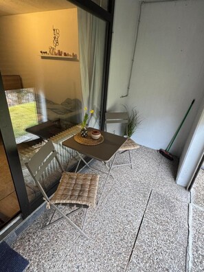 Un petit espace extérieur aménagé avec une table et des chaises, une fenêtre donnant sur un jardin et une porte menant à une autre pièce.