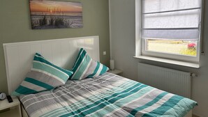 A bedroom with a bed, pillows, a nightstand, a wall clock, a painting, and a window with a view. - Apartment 'Meerestraum' with Wi-Fi (Norden)