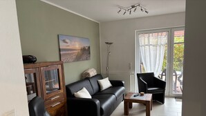 A living room with a sofa, chairs, a coffee table, a painting, a radiator, and a view through a glass door. - Apartment 'Meerestraum' with Wi-Fi (Norden)