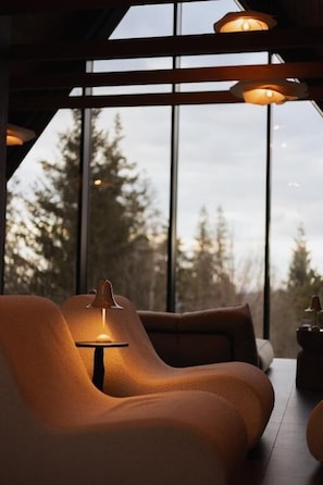 A modern interior with a sofa and a table lamp, large windows with a view of trees, and a ceiling with exposed beams.