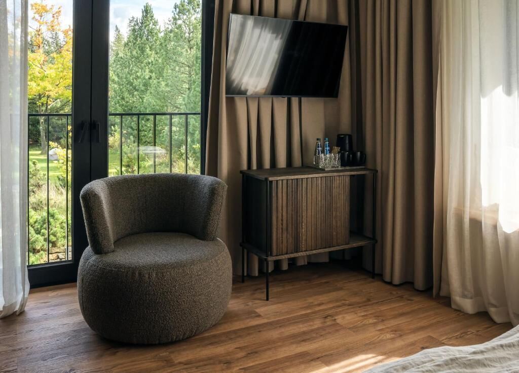 A room with a round chair, a TV mounted on the wall, a wooden cabinet with bottles, and a view of trees through a window.