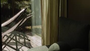 Una habitación con sofá, ventana con cortinas y vistas a una terraza de madera con hamaca.