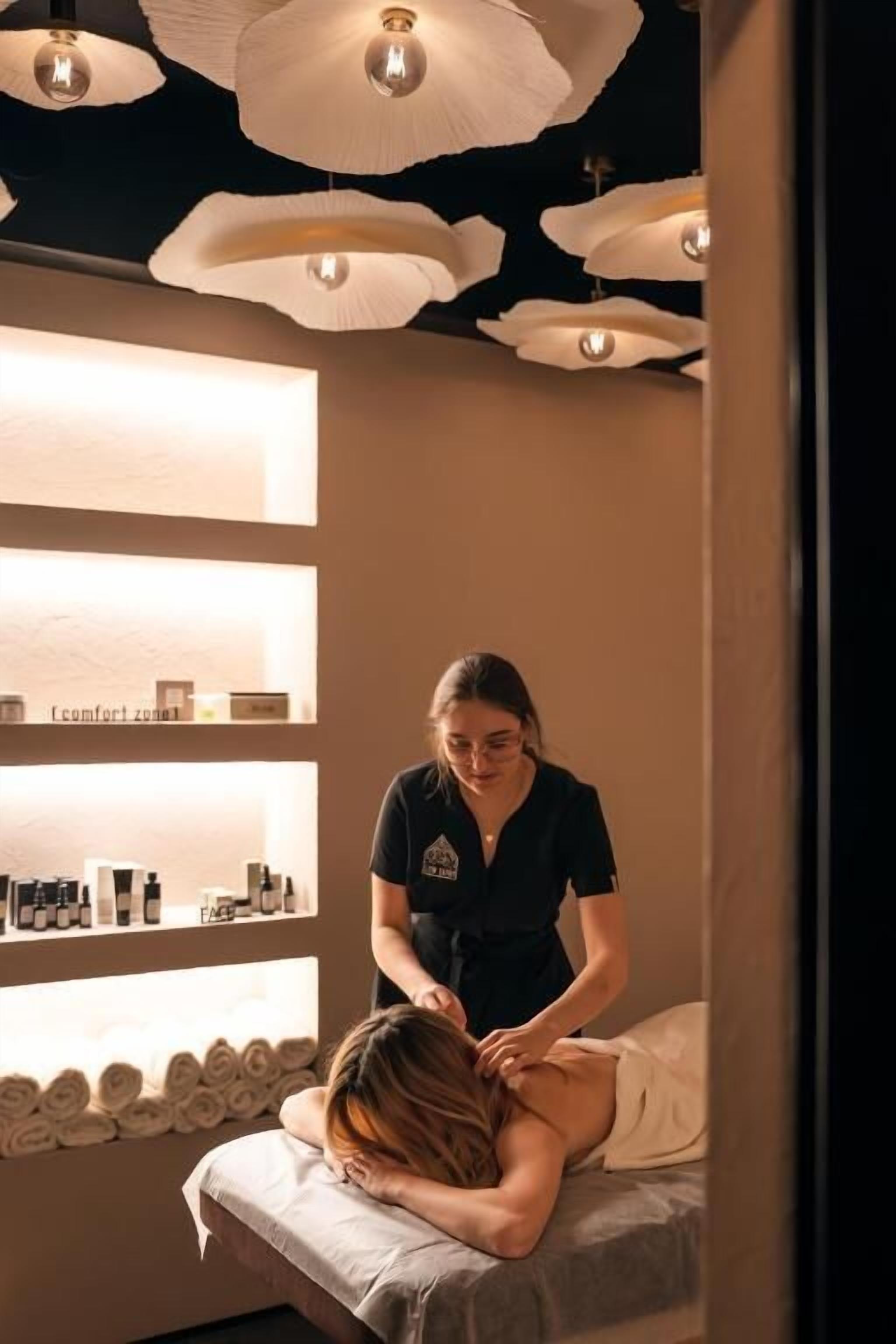 Une salle de massage équipée d’une table de massage, où un client reçoit un massage du dos, et des étagères remplies de produits de spa.