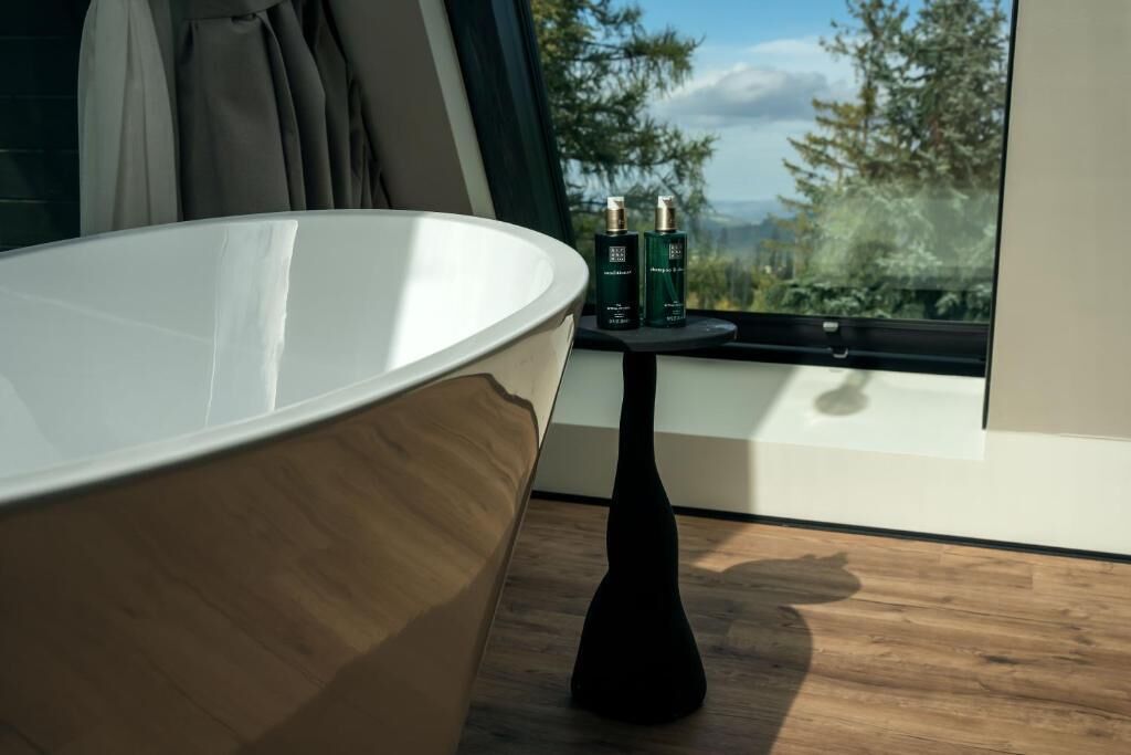 A modern bathroom with a large bathtub, a small table with bottles, and a view of trees through a window.