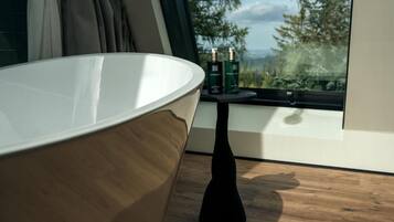 A modern bathroom with a large bathtub, a small table with bottles, and a view of trees through a window.