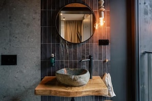 Une salle de bain avec un miroir rond, un lavabo en pierre, un plan de toilette en bois, un porte-serviettes et un luminaire fixé au mur.