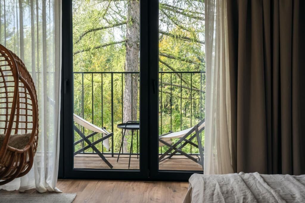 A room with a bed, a wicker chair, a balcony with a table and chairs, and a view of trees.