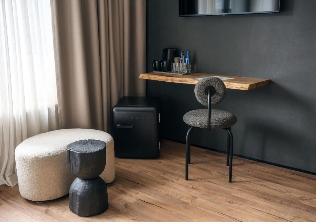 A modern room with a wooden floor, a black desk, a grey upholstered chair, a white ottoman, and a black suitcase.