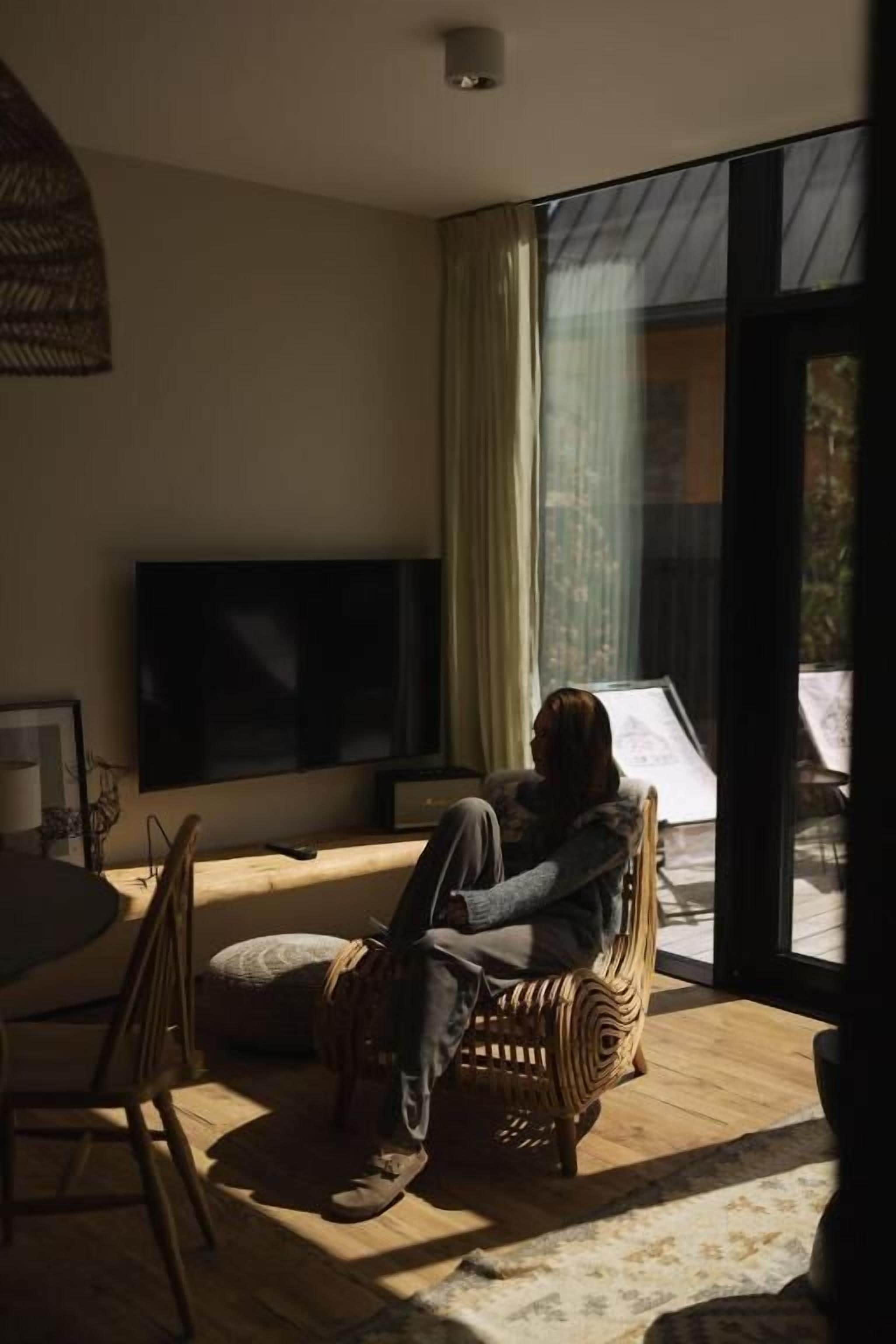 Una persona sentada en una silla de mimbre junto a un ventanal con vistas al exterior.