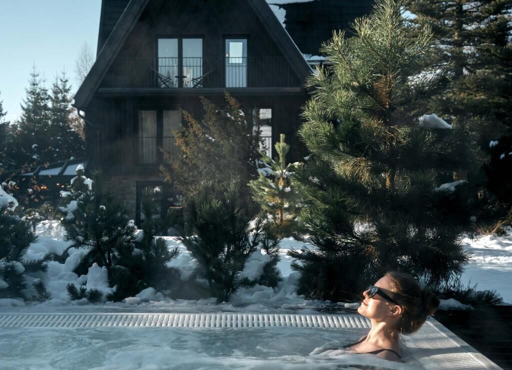 Una persona en un jacuzzi, con una casa y árboles cubiertos de nieve al fondo.
