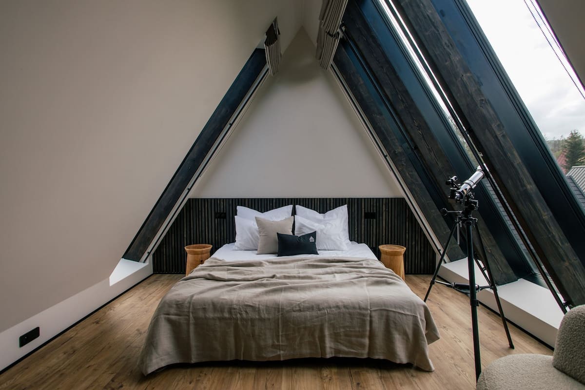 A bedroom with a sloped ceiling, a bed, a telescope, and a window.