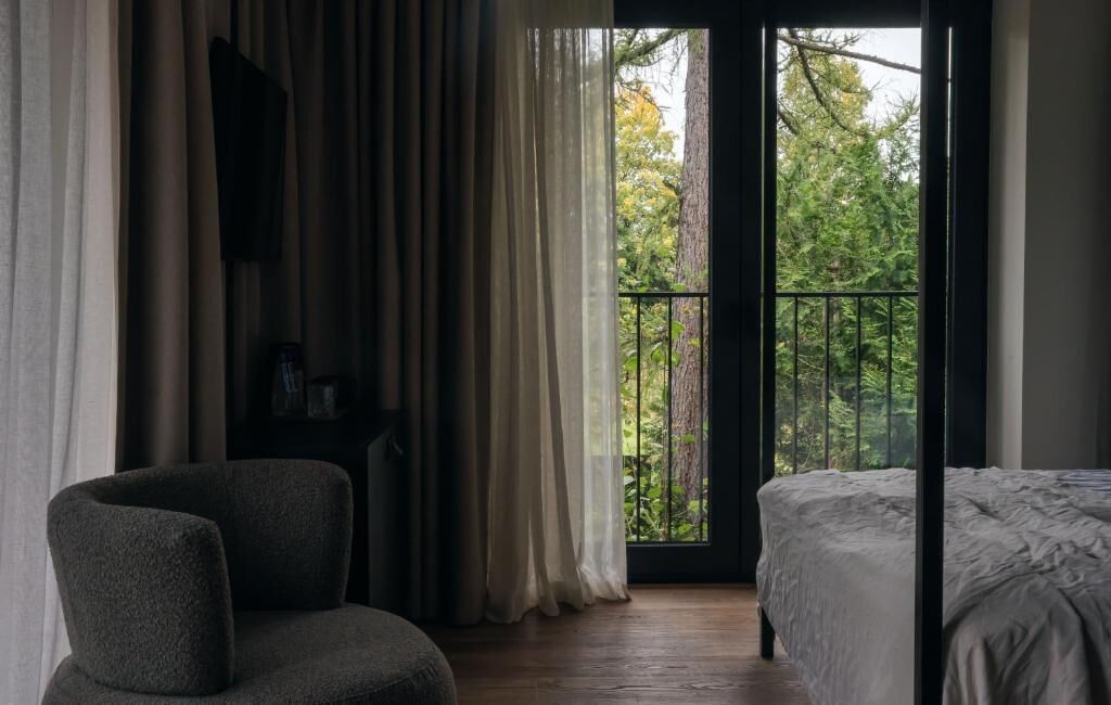 A bedroom with a bed, a chair, a desk, and a view of trees through a balcony door.