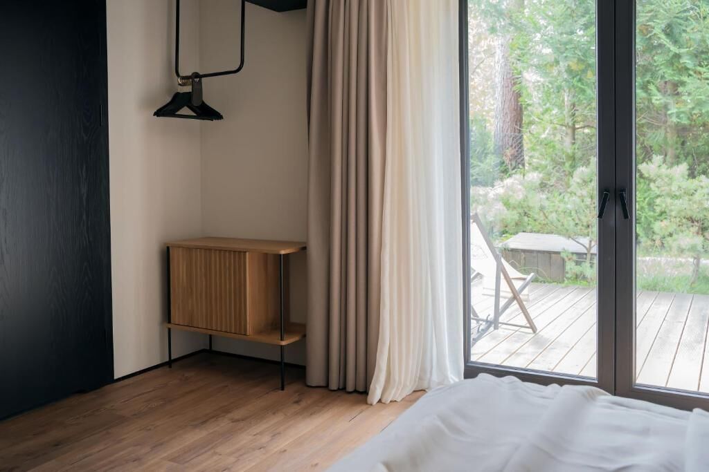 Un dormitorio con piso de madera, una cama, un mueble de madera pequeño, una ventana con cortinas y vistas a una terraza y vegetación.