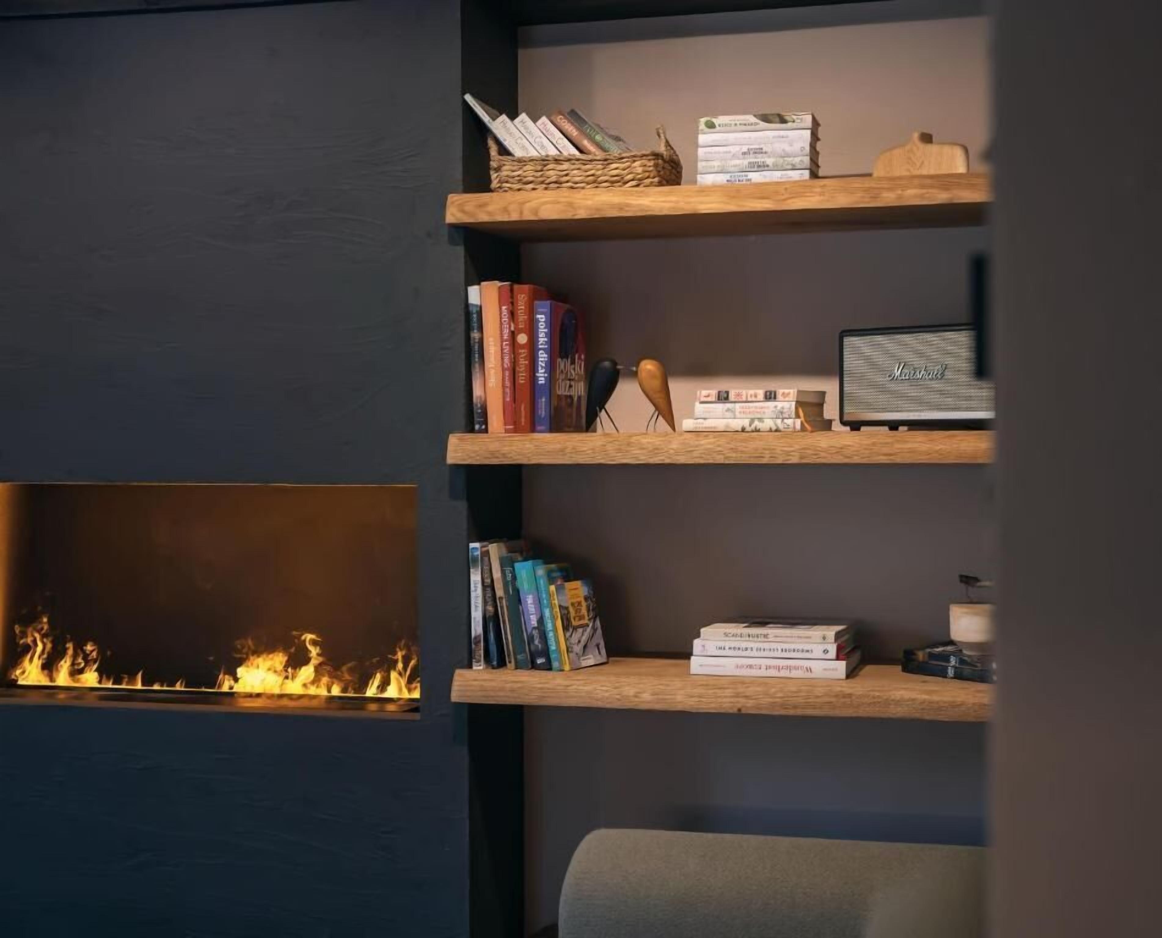 Uma estante com livros e objetos decorativos, uma lareira com chamas visíveis e uma parede escura.
