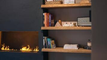 Uma estante com livros e objetos decorativos, uma lareira com chamas visíveis e uma parede escura.