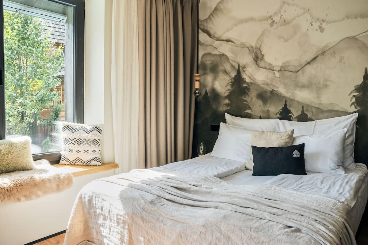 A bedroom with a bed, a window with curtains, a wall mural, and a small table with a lamp.